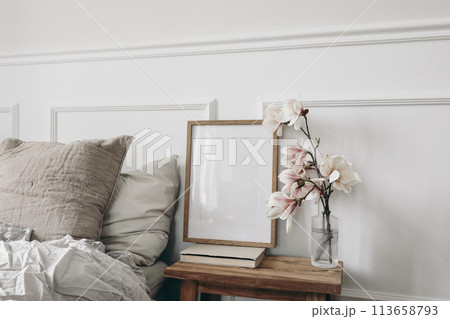 Empty vertical wooden picture frame mockup, old books. Wooden night stand with fluted glass vase. Blooming magnolia tree branches. Scandinavian interior, bedroom. White wall background, side view Empty vertical wooden picture frame mockup, old books. Wooden night stand with fluted glass vase. Blooming magnolia tree branches. Scandinavian interior, bedroom. White wall background, side view 113658793