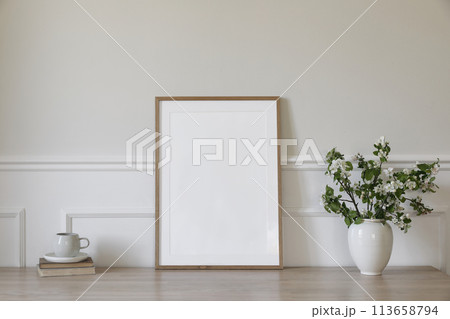 Springtime breakfast still life. Empty picture frame poster mockup. White ceramic vase with blooming apple tree branches. Cup of coffee, tea on wooden table, desk with old books. Scandi home interior Springtime breakfast still life. Empty picture frame poster mockup. White ceramic vase with blooming apple tree branches. Cup of coffee, tea on wooden table, desk with old books. Scandi home interior 113658794