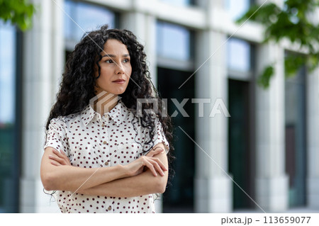 Portrait of a thoughtful Hispanic woman wearing a polka dot shirt, standing confidently outdoors with crossed arms near a modern building. 113659077