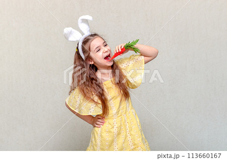 A girl with rabbit ears on her head in a yellow dress is gnawing on a carrot. Copy space A girl with rabbit ears on her head in a yellow dress is gnawing on a carrot. Copy space 113660167