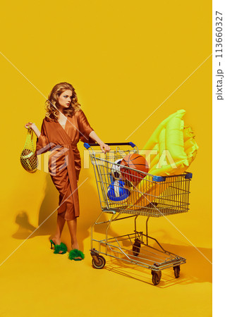 Woman in stylish brown dress pushes cart with colorful pool toys and sports equipment against yellow studio background. Woman in stylish brown dress pushes cart with colorful pool toys and sports equipment against yellow studio background. 113660327