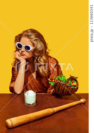 Woman in sunglasses holds hotdog in baseball glove, with bat at wooden desk, against yellow studio background. 113660341