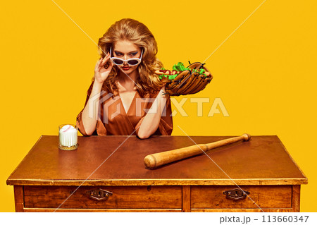 Stylish woman with sunglasses holds hotdog in baseball glove, with bat at wooden desk, against yellow studio background. 113660347