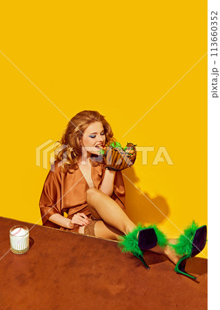 Woman with baseball glove eating hotdog with glass of milk stands on desk against yellow studio background. Woman with baseball glove eating hotdog with glass of milk stands on desk against yellow studio background. 113660352