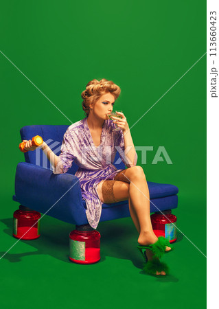 Girl drinks cocktail while sitting on vintage chair that has jars of vitamins instead of legs against vibrant green background. Girl drinks cocktail while sitting on vintage chair that has jars of vitamins instead of legs against vibrant green background. 113660423