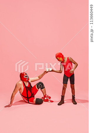 Masked wrestler in red gear seated, toasting milkshakes with standing partner against pink studio background. Masked wrestler in red gear seated, toasting milkshakes with standing partner against pink studio background. 113660449
