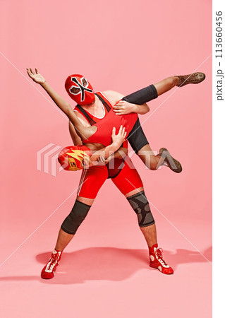 Athlete in red costume with mask executing wrestling move against partner dressed same costume against pink studio background. Athlete in red costume with mask executing wrestling move against partner dressed same costume against pink studio background. 113660456
