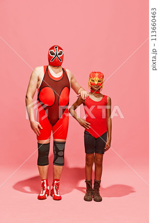 Two persons, middle-aged man and preteen boy, in superhero costumes posing against pink studio background. Two persons, middle-aged man and preteen boy, in superhero costumes posing against pink studio background. 113660463