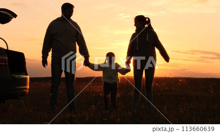 Family holding hands at sunset, silhouettes near car. Joint recreation in nature. Mother father daughter in orange setting sun in field. Family picnic road trip, parents kid enjoy each other's company 113660639