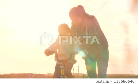 Silhouettes of child and mother, riding bike at dawn, sunset. Parent in field teaching educating girl skill of riding bicycle. Glare from sunrise sun, family summer fun. Mom training instructing kid. Silhouettes of child and mother, riding bike at dawn, sunset. Parent in field teaching educating girl skill of riding bicycle. Glare from sunrise sun, family summer fun. Mom training instructing kid. 113660640