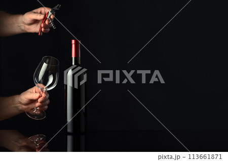 Bottle of red wine and hands with corkscrew and wine glass. 113661871
