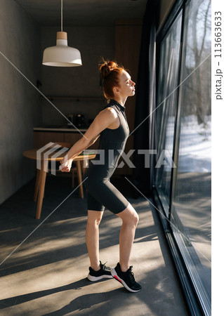 Side view of sporty slim young woman in outfit preforming warming up exercise, stretching ankle and arm muscles before workout, standing by window with beautiful snowy winter nature view on sunny day Side view of sporty slim young woman in outfit preforming warming up exercise, stretching ankle and arm muscles before workout, standing by window with beautiful snowy winter nature view on sunny day 113663633