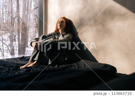Portrait of sad young woman sitting by window on sunny day on windowsill with winter nature background, feels upset, having melancholic mood, thinking about life troubles. Loneliness concept. 113663710