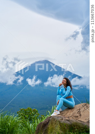 A young woman sits on a large rock in a viewpoint and enjoys the view of the sacred Mount Agung volcano hidden by clouds on a rainy day on the island of Bali. 113667036