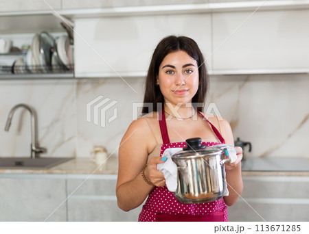 Young woman in apron posing with saucepan in kitchen 113671285
