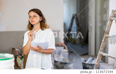 Portrait of positive woman next to a stepladder with documents in her hands checks the quality of the completed repairs in cottage room Portrait of positive woman next to a stepladder with documents in her hands checks the quality of the completed repairs in cottage room 113671594