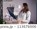 A young adult white female doctor in a medical mask and a white coat holds a CT scan in her hands and looks at it carefully 113676006