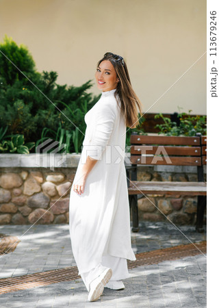 Happy woman in a white dress stands in front of a bench at city 113679246