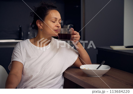 Young pretty woman in white pajamas, sitting with her eyes closed at the kitchen table, taking sip of a freshly brewed espresso coffee. Start your day with energizing freshly brewed coffee 113684220