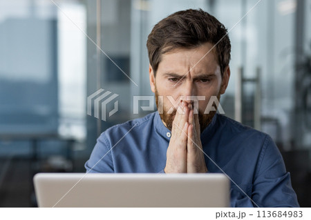 Shocked and worried young male businessman sitting in the office at his desk and looking thoughtfully at the laptop screen with his hands folded together. Shocked and worried young male businessman sitting in the office at his desk and looking thoughtfully at the laptop screen with his hands folded together. 113684983