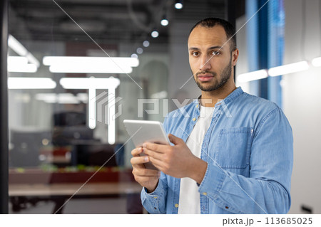 Portrait of a serious young Latin American man standing in the office, holding a tablet and looking at the camera. 113685025