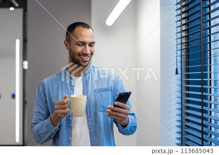 Smiling young arab man working in office, standing by window on work break, holding cup with drink and using mobile phone. Smiling young arab man working in office, standing by window on work break, holding cup with drink and using mobile phone. 113685043