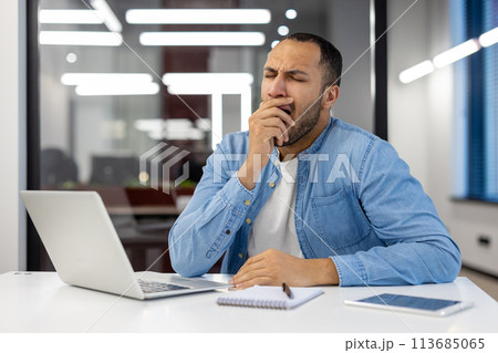 A tired young Muslim man in a blue shirt sits at a desk in a modern office and yawns while covering his mouth with his hand. A tired young Muslim man in a blue shirt sits at a desk in a modern office and yawns while covering his mouth with his hand. 113685065