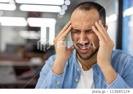 A close-up photo of a Muslim young man sitting in the office behind the workplace and hunched over from anger and tension, holding his head in his hands. 113685124