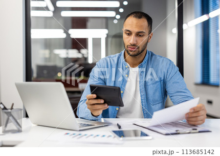 Serious young hispanic male accountant and auditor sitting in office at desk working with documents, bills and calculator. 113685142