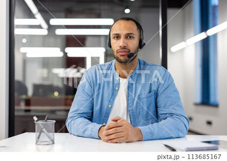 Portrait of a serious Muslim man sitting at a desk in the office, wearing a headset, confidently looking at the camera. 113685167