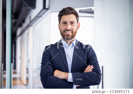 Portrait of a smiling and successful young businessman standing in the office, crossing his arms on his chest and looking at the camera. 113685171