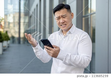 Angry and worried young Asian male businessman standing outside a building, looking upset at the phone screen and throwing up his hands. Angry and worried young Asian male businessman standing outside a building, looking upset at the phone screen and throwing up his hands. 113685239