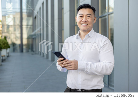 Portrait of a smiling young Asian businessman in a white shirt standing outside, holding a mobile phone and looking at the camera. 113686058