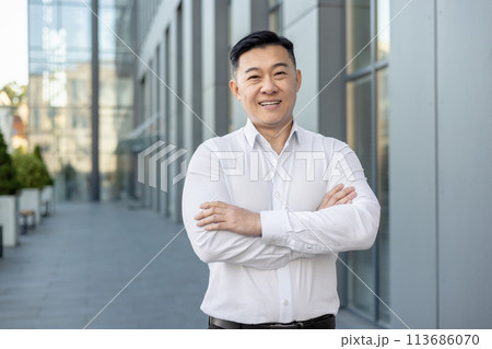 Portrait of a smiling and successful young businessman, office worker in a white shirt standing outside on the street, crossing his arms on his chest and looking confidently at the camera. Portrait of a smiling and successful young businessman, office worker in a white shirt standing outside on the street, crossing his arms on his chest and looking confidently at the camera. 113686070