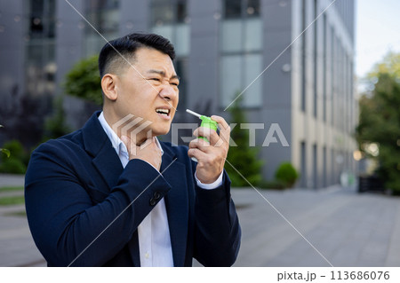 Asian young businessman sitting near working office outside, holding throat with hand, feeling severe pain and using medical spray. Asian young businessman sitting near working office outside, holding throat with hand, feeling severe pain and using medical spray. 113686076