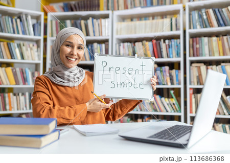 A cheerful female educator in a hijab presenting the concept of 'Present Simple' on a whiteboard in a library, surrounded by books. A cheerful female educator in a hijab presenting the concept of 'Present Simple' on a whiteboard in a library, surrounded by books. 113686368