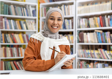 A cheerful young Muslim woman wearing a hijab studies from her notebook in a library, smiling as she looks towards the camera, surrounded by bookshelves. 113686401
