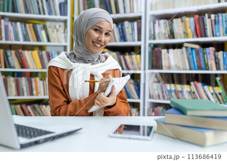 A cheerful young woman in a hijab sits at a library desk, studying from notes and using a laptop, surrounded by books. Represents focus and academic commitment. 113686419