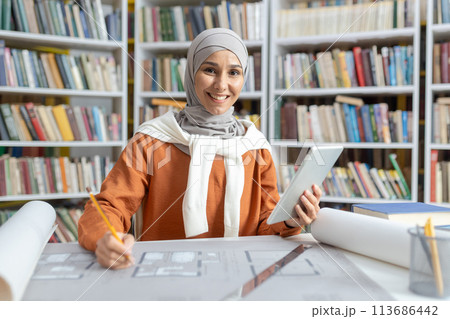 Confident Muslim female architect working on project plans with a digital tablet in a library surrounded by books. She is wearing a hijab and a bright orange top, engaging warmly with the camera. Confident Muslim female architect working on project plans with a digital tablet in a library surrounded by books. She is wearing a hijab and a bright orange top, engaging warmly with the camera. 113686442