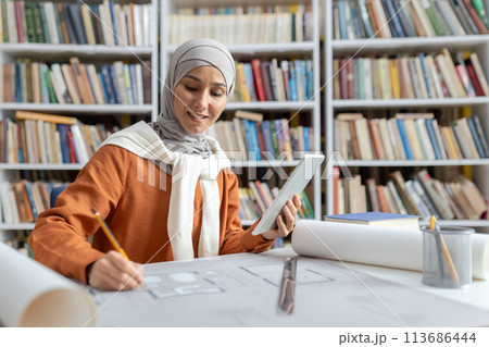 A focused Muslim woman architect, dressed in a hijab, reviews digital blueprints on a tablet while working with physical drafts in a well-stocked library. 113686444
