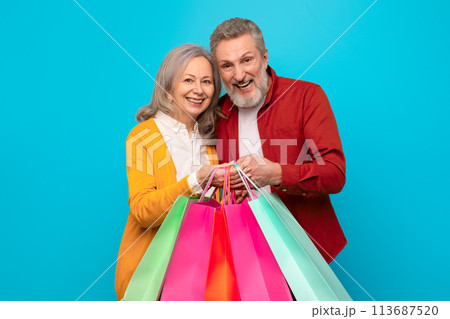 Cheerful retired mature couple holding many shopper bags, blue backdrop 113687520