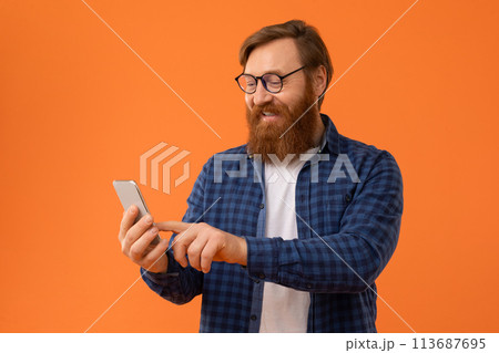 redhaired Caucasian man with beard uses his smartphone, orange backdrop redhaired Caucasian man with beard uses his smartphone, orange backdrop 113687695