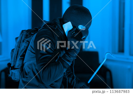 Burglar with phone and crowbar at night Burglar with phone and crowbar at night 113687983