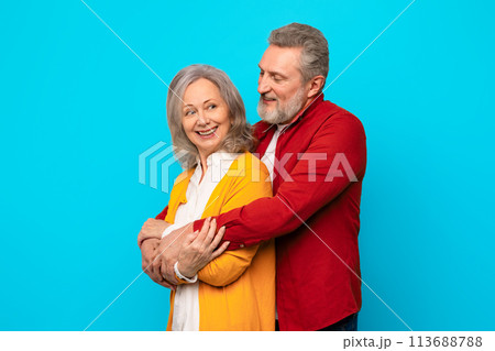 Happy loving senior couple embracing with affection against blue backdrop Happy loving senior couple embracing with affection against blue backdrop 113688788