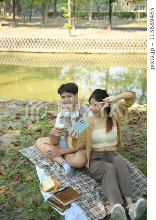 Cheerful young couple sitting on on blanket in park and taking selfie with smartphone Cheerful young couple sitting on on blanket in park and taking selfie with smartphone 113689465