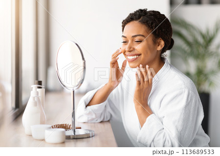 Happy young black woman applying facial moisturizer while looking in makeup mirror Happy young black woman applying facial moisturizer while looking in makeup mirror 113689533