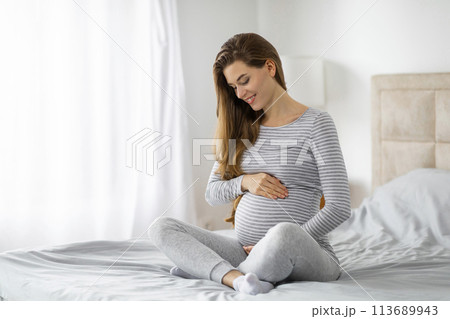 Pregnant woman sitting on bed holding belly Pregnant woman sitting on bed holding belly 113689943