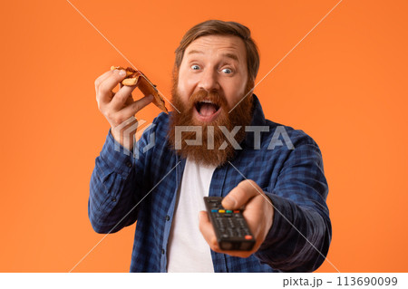 redhaired bearded guy points remote controller while enjoying pizza, studio 113690099