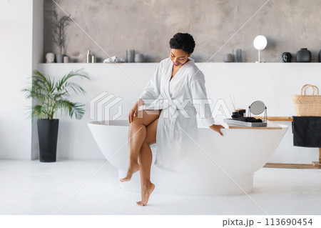 Black woman wearing bathrobe sitting on bathtub and touching her beautiful legs 113690454