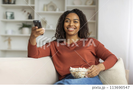 Smiling Black Woman Watching TV and Holding Remote at Home 113690547